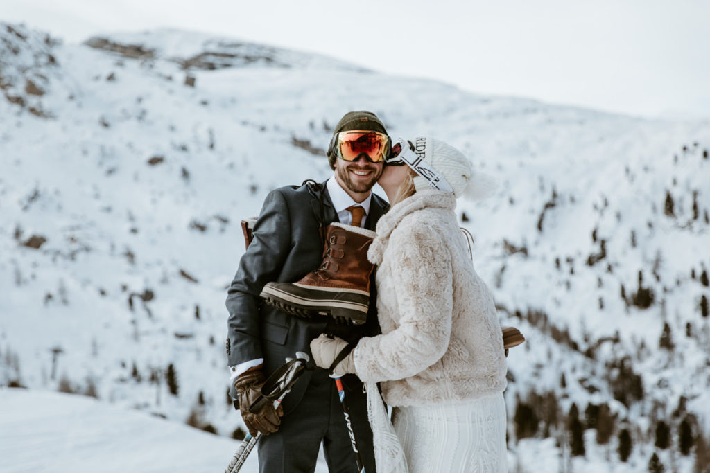 Couple stands on the slopes during their two day Dolomites ski wedding. He is wearing ski goggles with his hiking boots over his shoulders and she is sideways holding ski poles, kissing the side of his face.