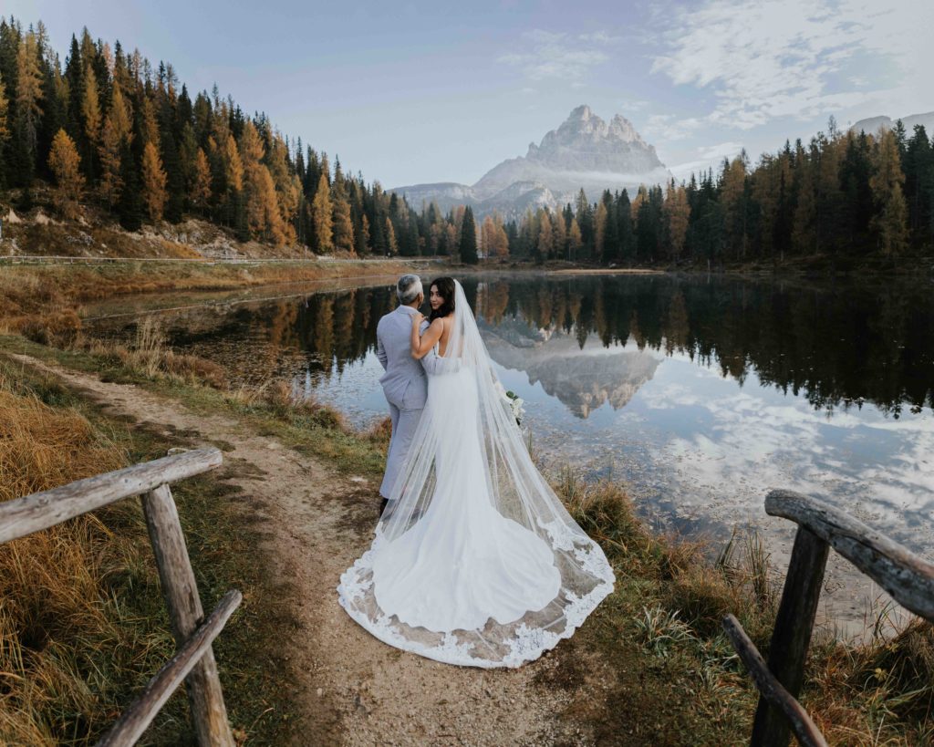 A couple stands near lago d'Antorno in the Dolomties during their fall elopement. The lake is still and the mountains are reflected. The leaves are golden and the woman is looking back over her shoulder at the camera.