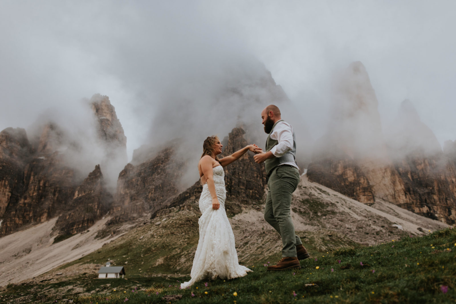 A couple stand under the Tre Cime peaks in the Dolomites during their spring elopement. There is dramatic fog between the peaks and they are holding hands and dancing.