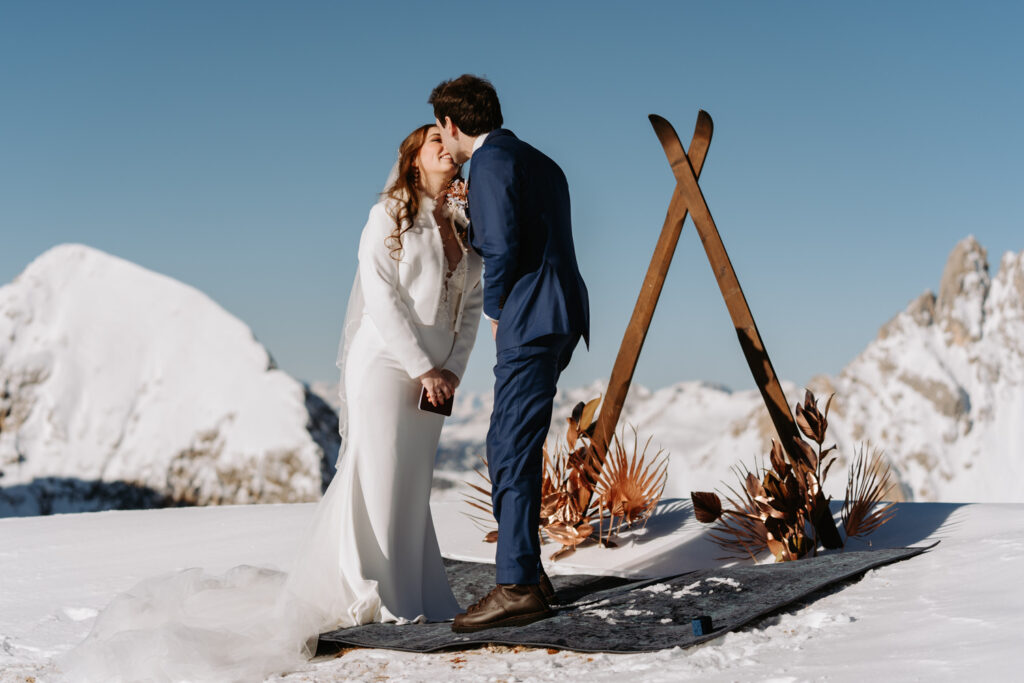 A couple in white wedding dress and blue suit lean in for a kiss atop a snow covered mountain surrounded by skis and flowers on their Dolomites ski elopement day