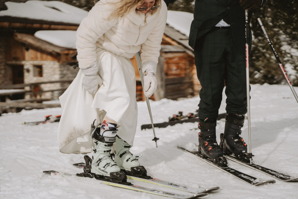 A couple in wedding clothes puts on their skis in the Dolomites during their winter ski elopement