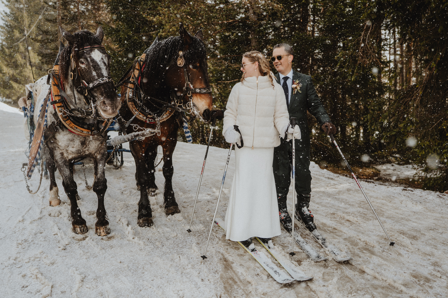 A couple on skis wearing winter wedding attire stand next to horses in the Italian Dolomites for their elopement. They are standing facing the horses and smiling, as fat snowflake fall around them.