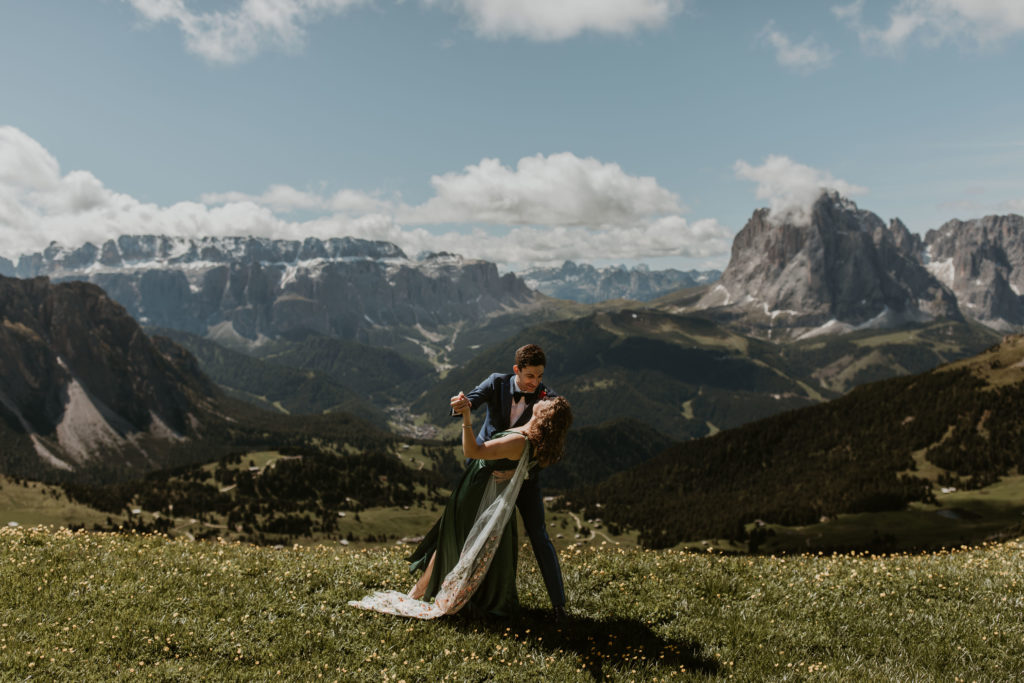 A couple stands wearing wedding attire, doing a dip near Seceda in the Italian Dolomites. The grass is very green and there are small yellow flowers. The sun is shining.
