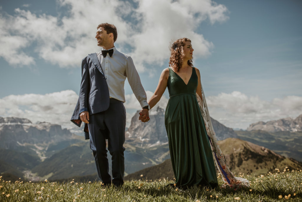 A couple eloping in the Dolomites stands holding hands and looking in opposite directions on a grassy mountain field.