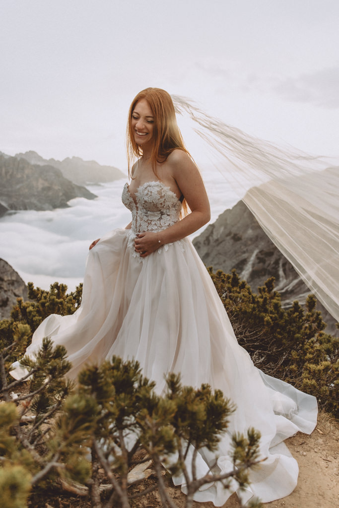 This is a photo of a bride during her elopement in the Dolomites. She is holding her dress and laughing. There are dramatic clouds in the background.
