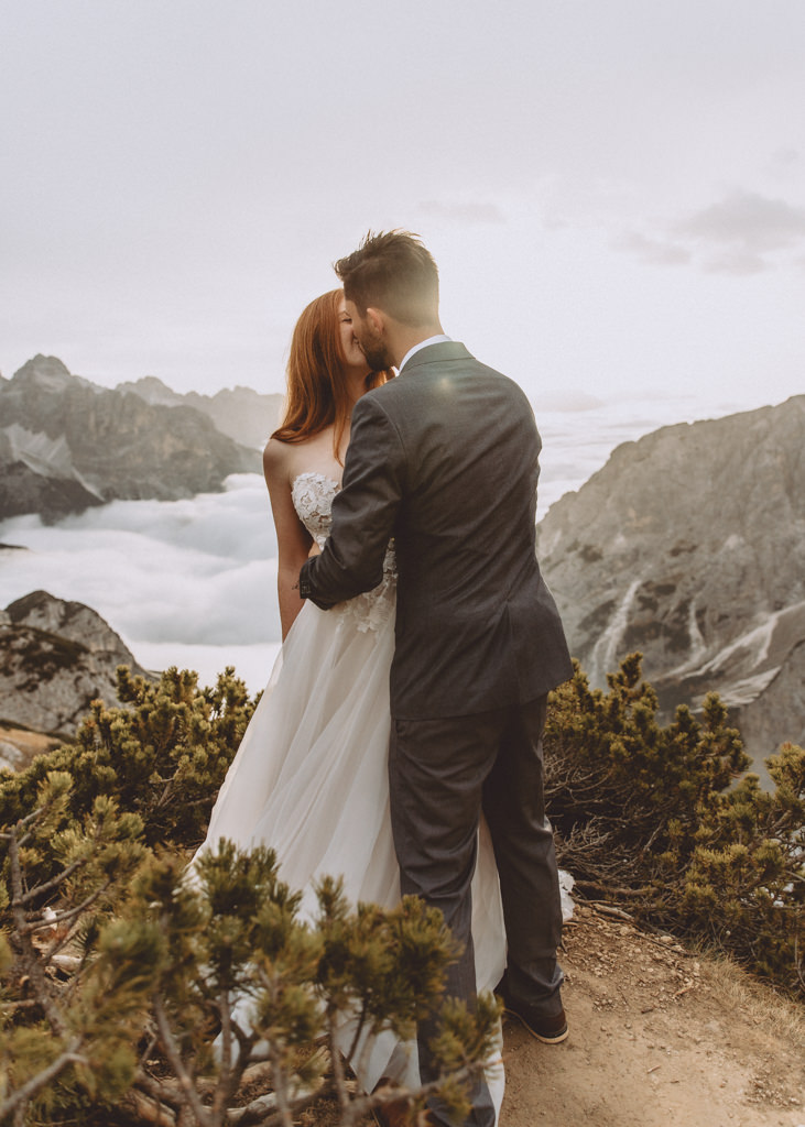 This is a photo of a spring elopement in the Dolomites. The couple is kissing and there is dramatic fog below them and vast mountain views.