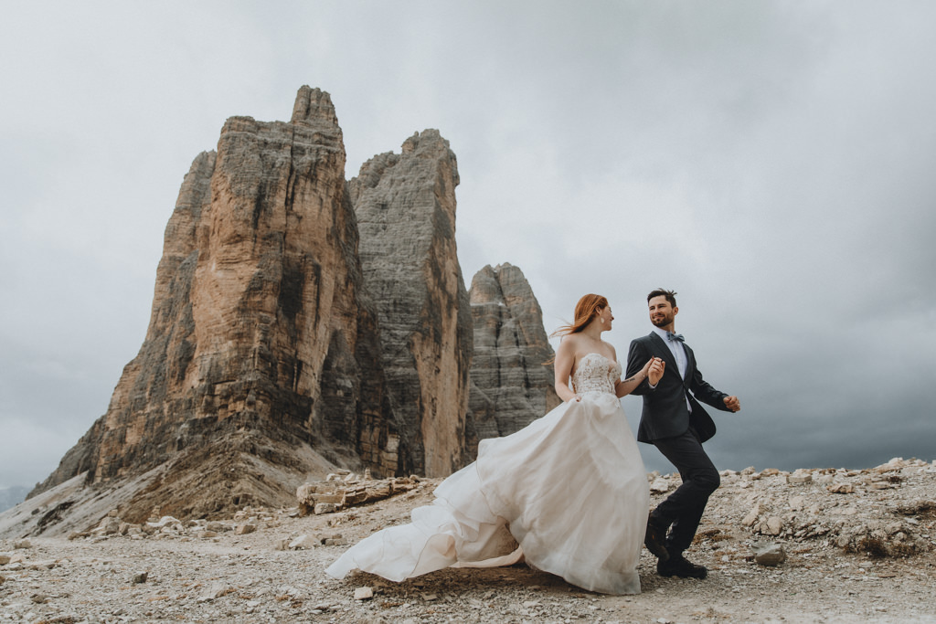A couple wearing wedding clothes is running on a rocky trail in the Italian Dolomites. They are looking at each other. The Tre Cime mountain peaks on in the background, and dark clouds stretch in the distance.