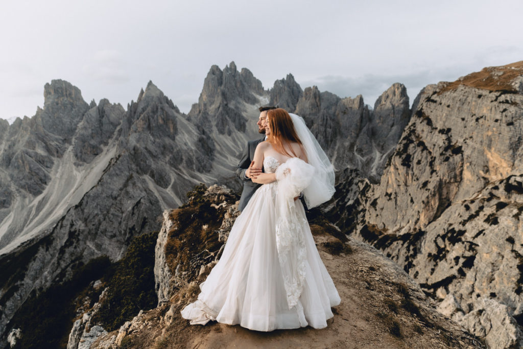 This is a photo of a spring elopement photos at Cadini di Misurina in the Dolomites. The couple is standing looking to the left toward the light, smiling, with dramatic mountain peaks behind them.