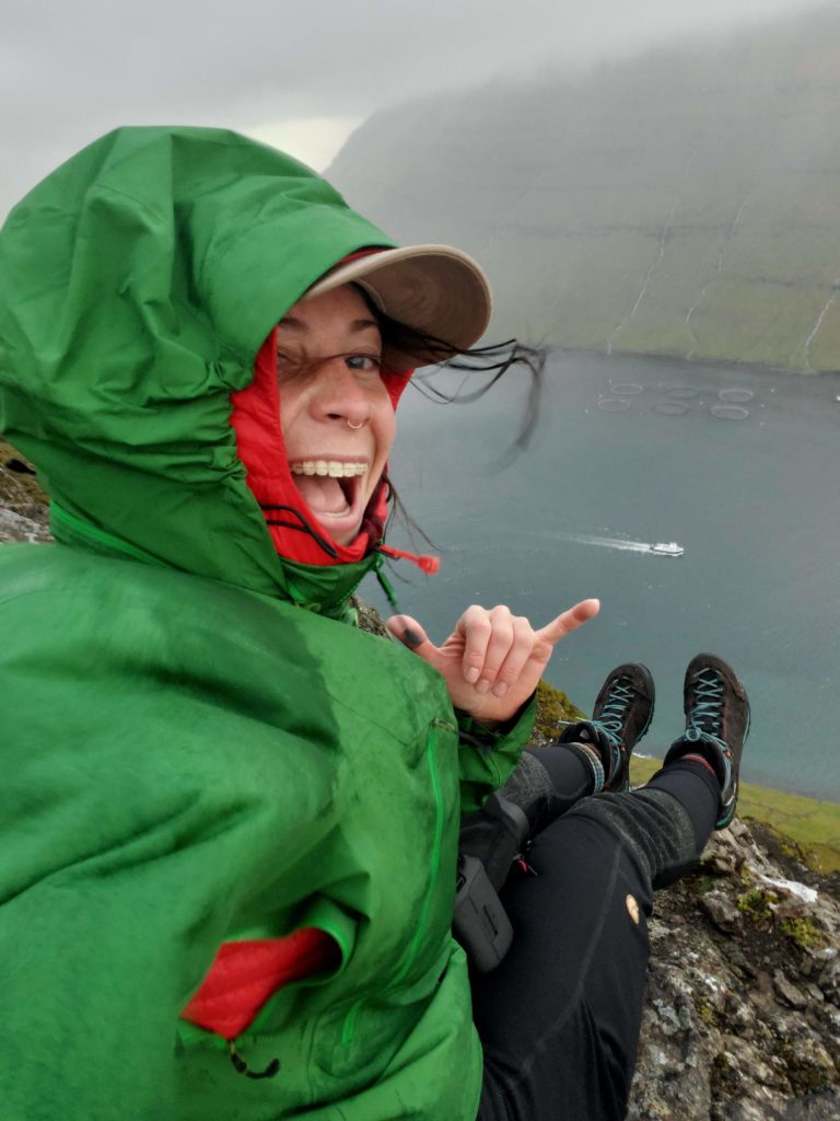 This is a photo of the hiking trail Klakkur on the Faroe islands. There is a woman in a green rain jacket sitting on the edge of a cliff overlooking the fjord. There is a small boat in the distance and she is smiling at the camera.