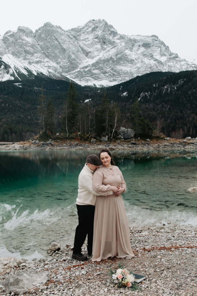 This is an image of couple during their Eibsee sunrise elopement. They are standing near the water embracing. The mountains are reflected in the background in the greenish water.