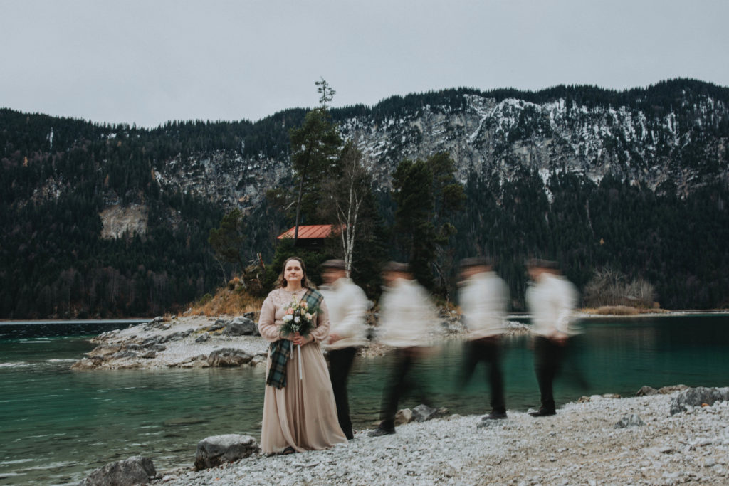 This is an image of a couple eloping at Eibsee. The woman is standing on a stone beach, facing toward the camera, holding a bouquet and wearing a long, light pink dress and sweater. The husband is shown walking in slow motion toward her, leaving a blurred light trail behind him. 