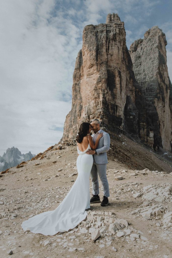 A couple stands forehead to forehead in their wedding attire under the Tre Cime peaks during their Dolomites hiking elopement. The peaks tower above them in the background.