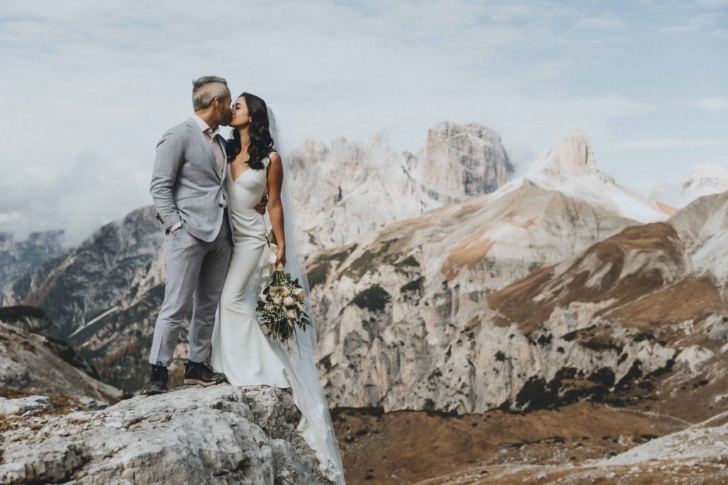 This is an image of a wedding couple standing on a boulder during their two day Dolomites hiking elopement. They are kissing and the bride is holding her bouquet while the groom holds her waist with one hand, his other in his pocket. There is a dramatic view of rocky mountains behind them.