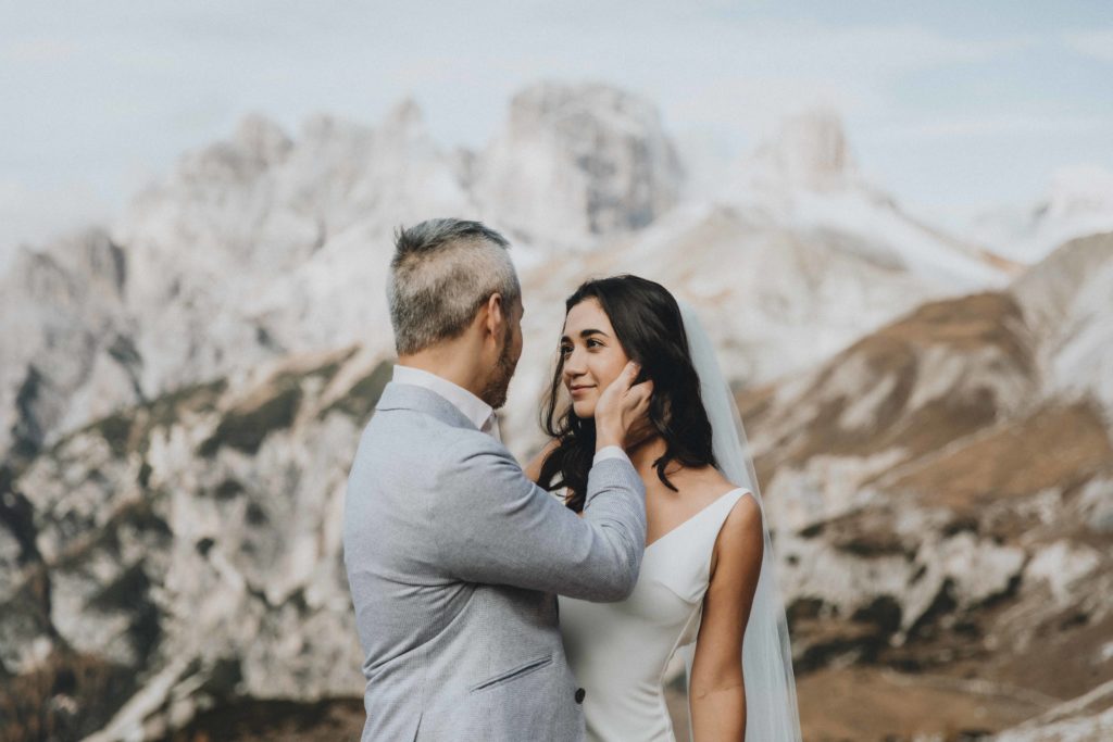 This is an image of a wedding couple standing on a boulder during their two day Dolomites hiking elopement. The groom is caressing her cheek while she smile at him. There is a dramatic view of rocky mountains behind them.