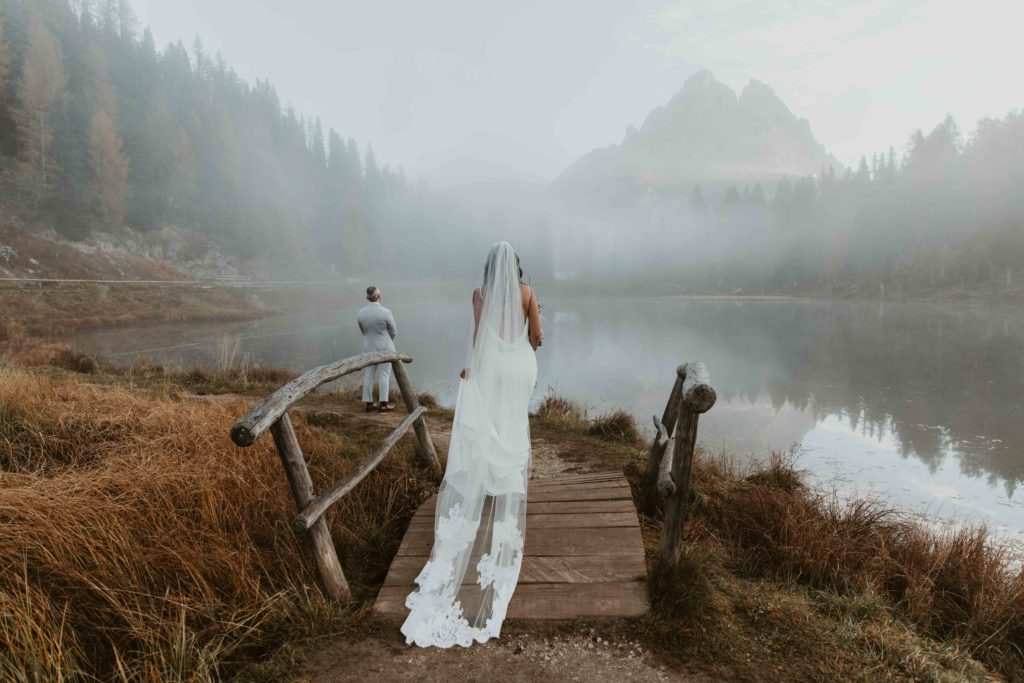 A woman in a wedding dress with a long train walks across a small wooden bridge to her groom, starting off into the mountain fog as he waits for his first look at her.
