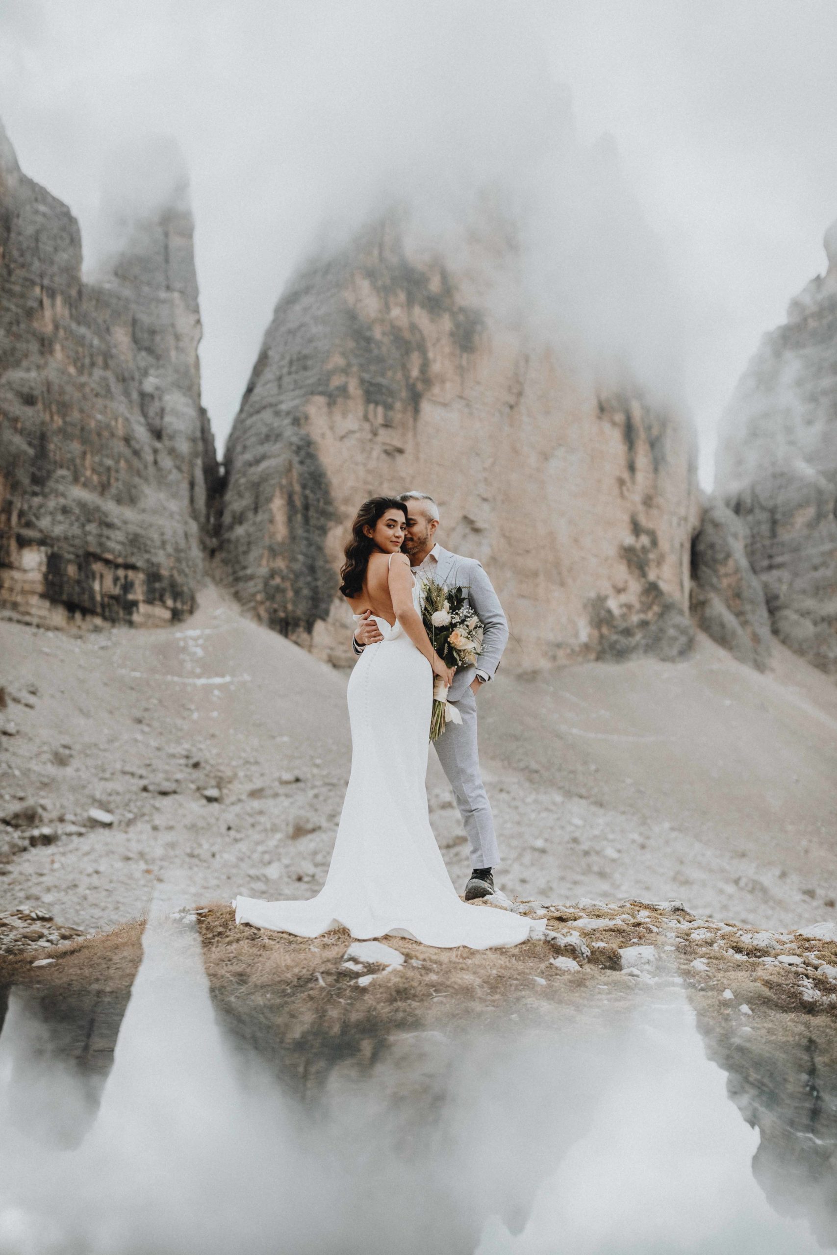 A couple stands cheek to cheek  in their wedding attire next to the Tre Cime peaks during their Dolomites hiking elopement. They peaks are covered in mist, and the woman is looking back at the camera dramatically.