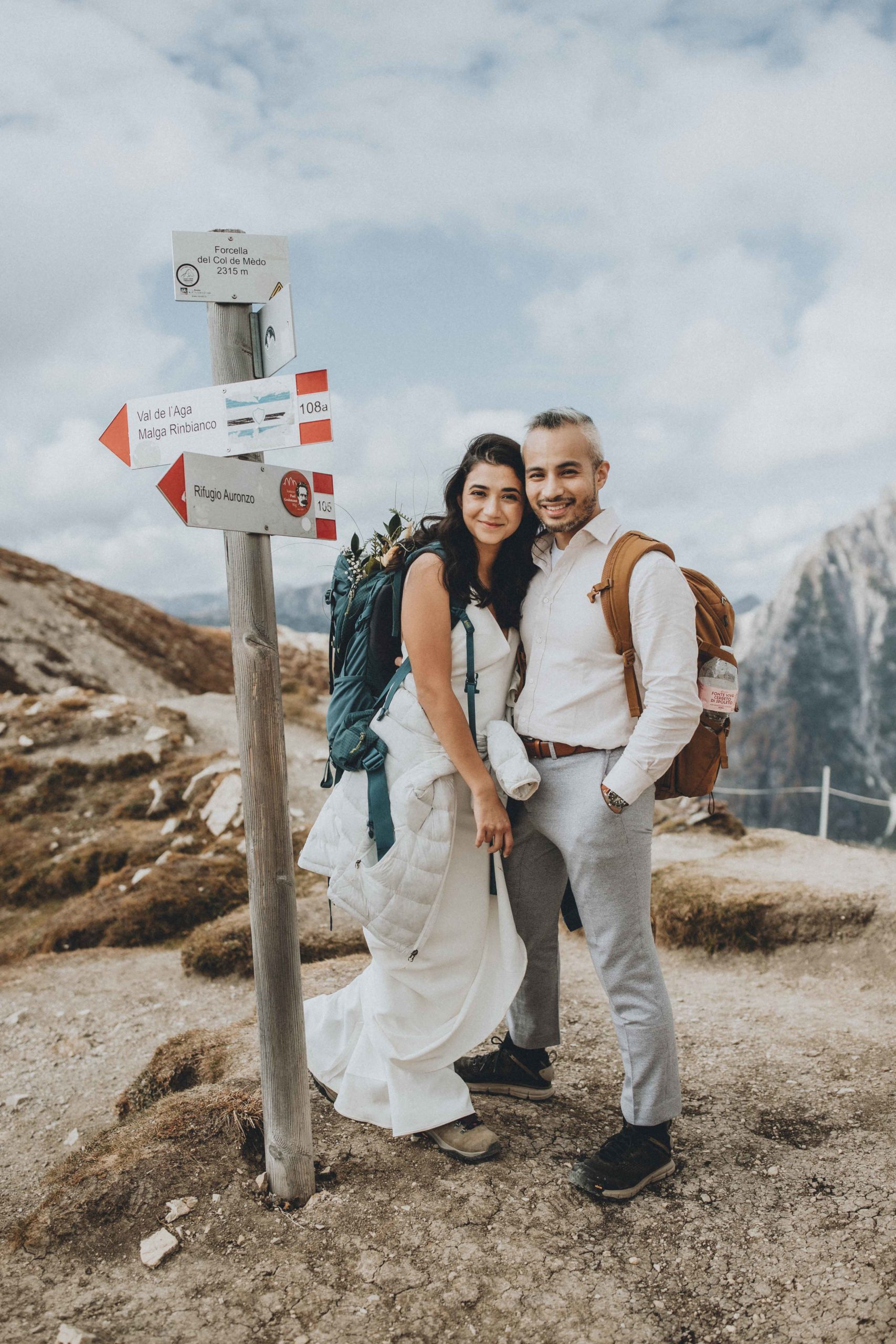This is an image of a couple eloping in the dolomites. They are standing in their wedding outfits next to a trail sign, smiling at the camera. They are wearing backpacks and hiking boots over their wedding outfits. 