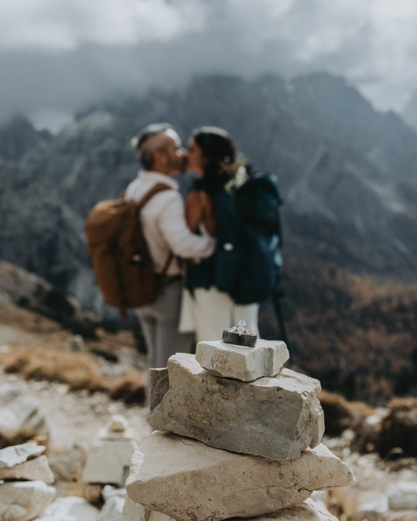 This is a photo of a couple during their Dolomites hiking elopement. They are out of focus in the background, kissing, while their wedding rings sit on a cairn in the foreground.