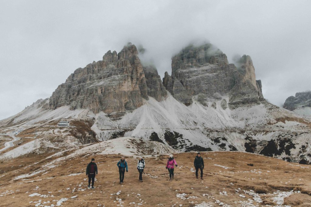A group of five people hike side by side toward the camera in the Dolomites with dramatic misty mountain peaks behind them