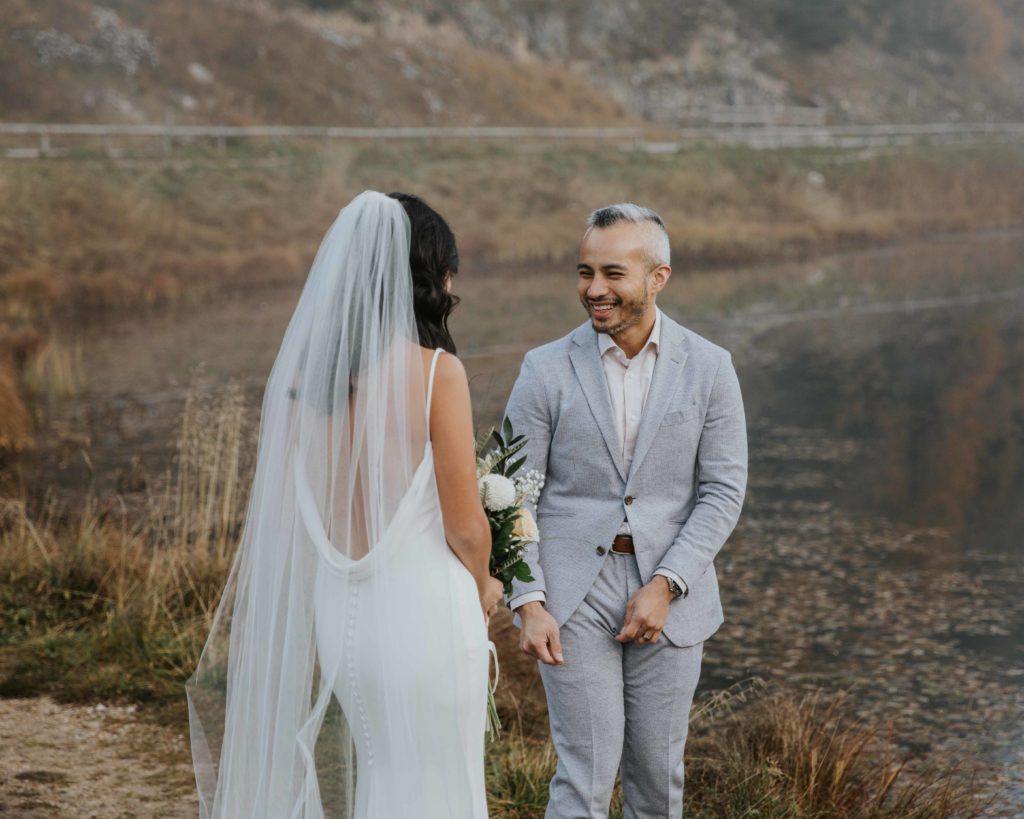 A groom wearing a light blue suit turns around with a big smile to see his bride for the first time during their elopement first look.