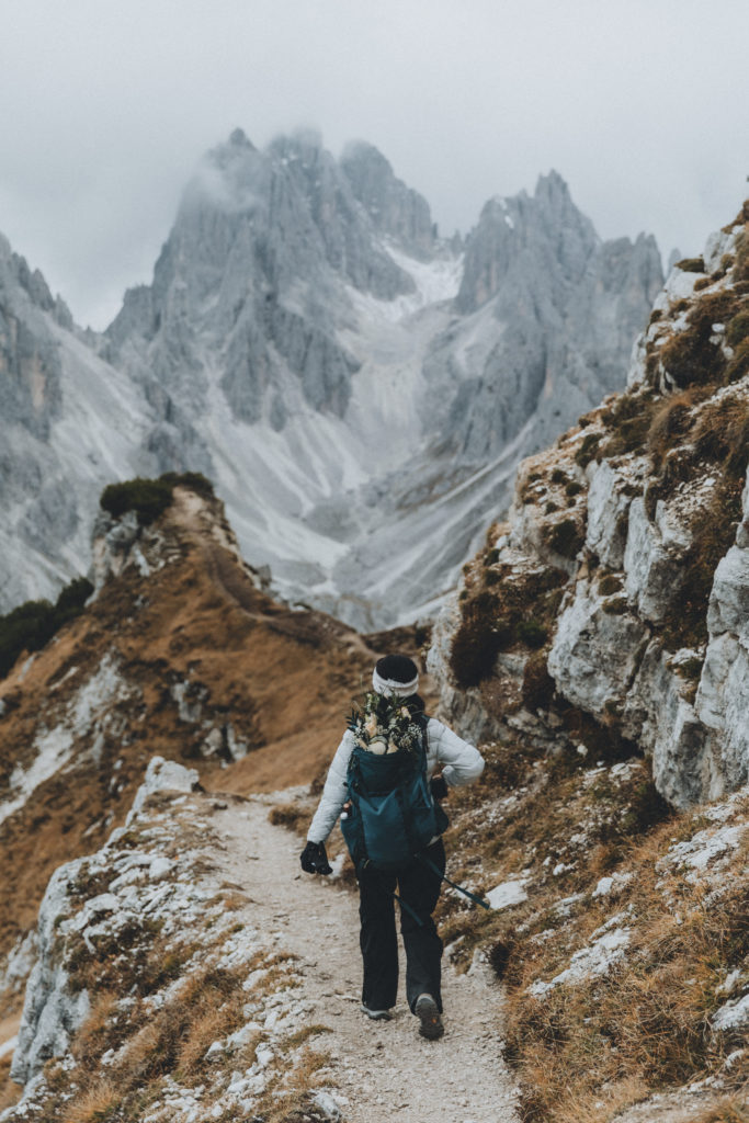 This is a photo of a woman hiking the Cadini di Misurina viewpoint trail in the Dolomites during her elopement. She is walking away from the camera toward dramatic peaks, wearing a white jacket and hat and hiking backpack.