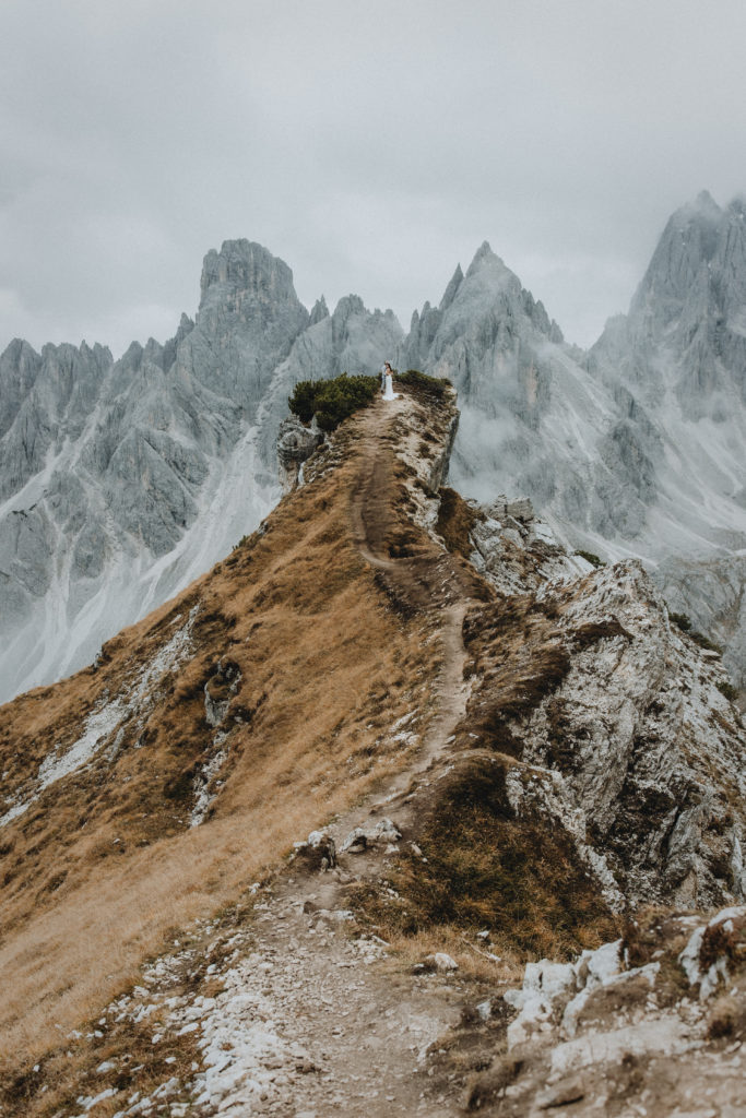 This is an image of a couple walking out on a narrow trail in the Italian Dolomites. They are very small in the frame and there are rocky, dramatic mountain ridges behind them.