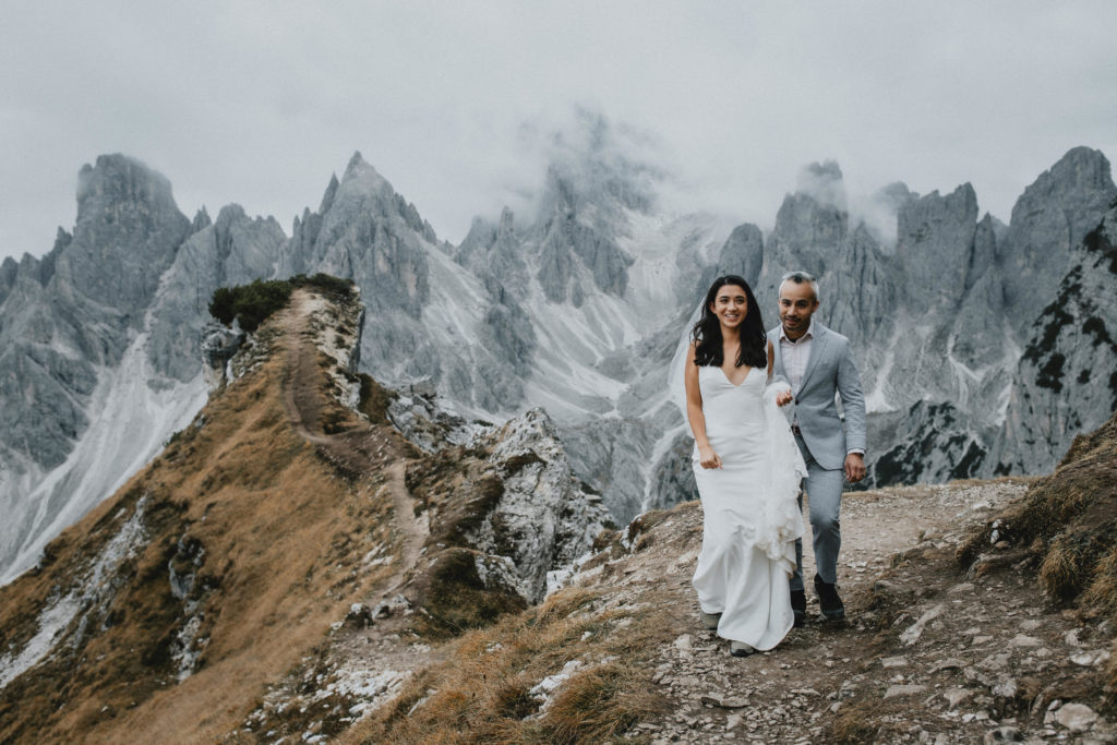 This is a photo of a wedding couple walking on the Cadini di Misurina viewpoint trail. They are hiking for their Dolomites elopement, smiling toward the camera with dramatic rocky peaks in the background.