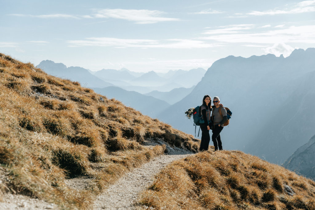 This is a photo of a couple standing on a grassy golden ridge, facing the camera, with blue mountains spreading into the distance. They are hiking for their Dolomites elopement.