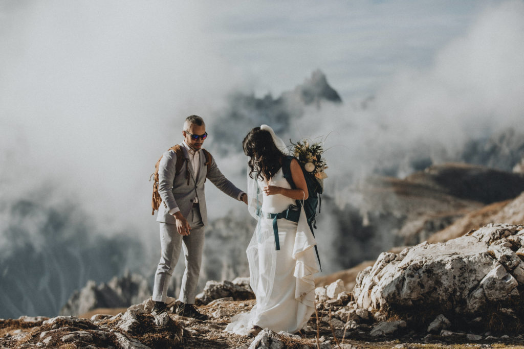 A couple stands on a misty mountaintop during their 2 day Dolomites hiking elopement. They are wearing their wedding gear and hiking packs, looking away from the camera.