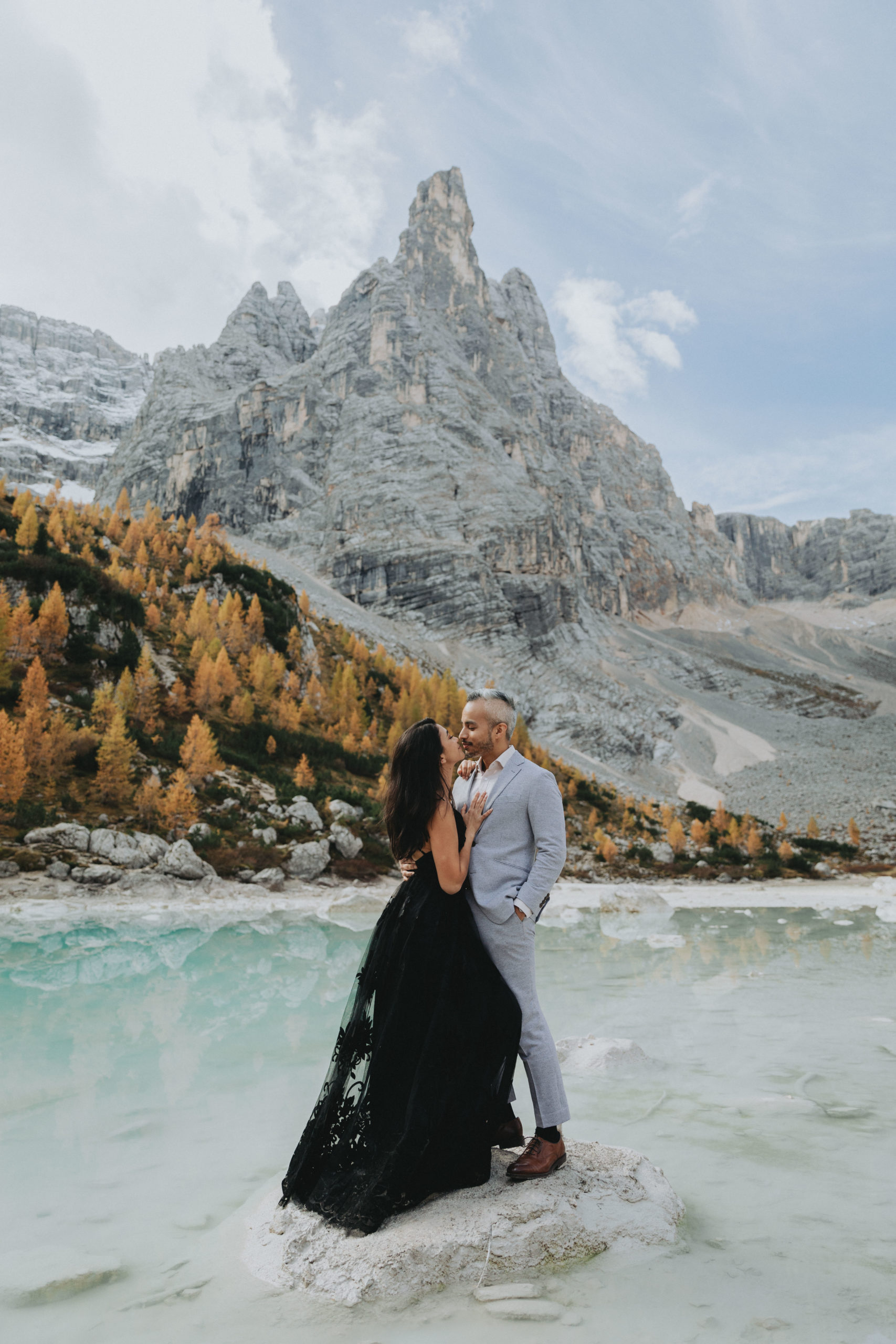 A couple embraces while wearing their wedding attire near the impressive Lago di Sorapis during their Dolomites elopement. The wife is wearing a black dress and he is wearing a light grey suit. They are standing nose to nose.