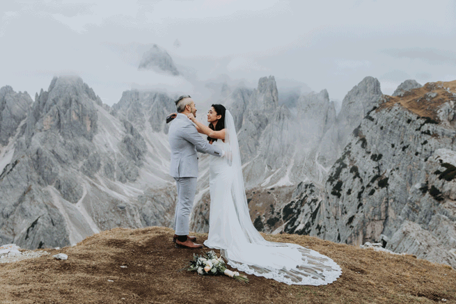 This is a gif of a couple dressed in their wedding gear going in for a kiss. They are standing on the top of a mountain in the Italian Dolomites during their elopement. The peaks are grey and jagged behind them, covered in clouds. 