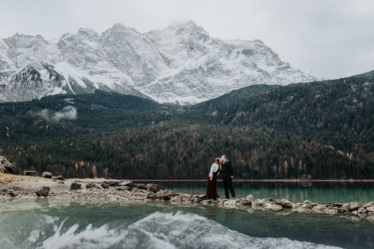 This is an image of a couple eloping at Eibsee during sunrise. They are standing on a narrow path of stones across the lake, kissing. The mountains are tall and snow covered in the background, reflected in the greenish lake.