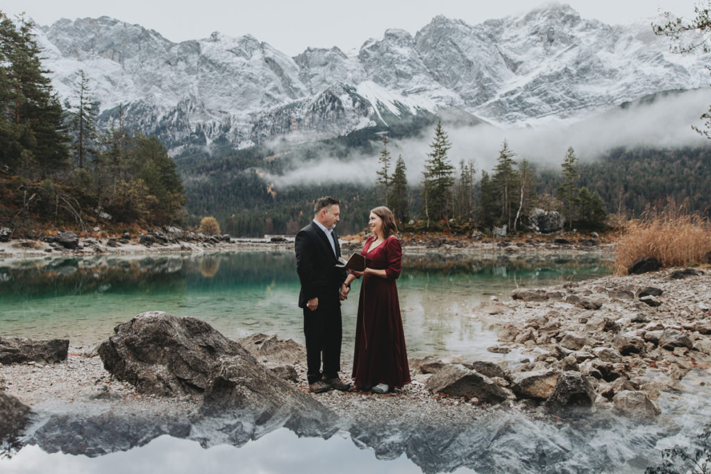 This is a photo of couple eloping at Eibsee during sunrise. They are standing, facing each other, near a deep green lake surrounding by trees and tall mountain peaks in the background. The mountains are misty. They are holding hands.
