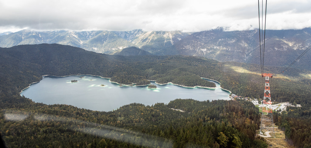 This is an image of the view over Eibsee from the Zugspitze cable car. There is an oblong, green lake dotted with small islands and surrounded by thick evergreen trees and mountains. The cable car wires stretch across the right side of the frame.
