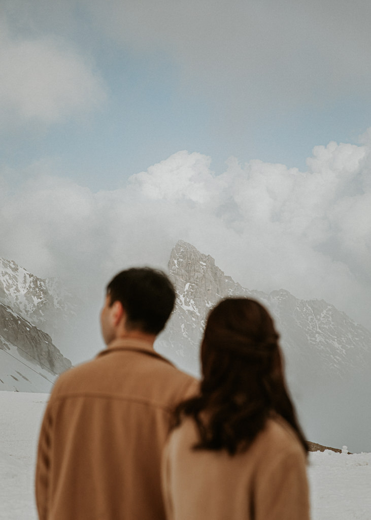This is an image of a couple having their photo taken at the top of the Zugspitze, near Eibsee. The mountain peak is in focus in front of them. They are facing away from the camera, slightly out of focus.