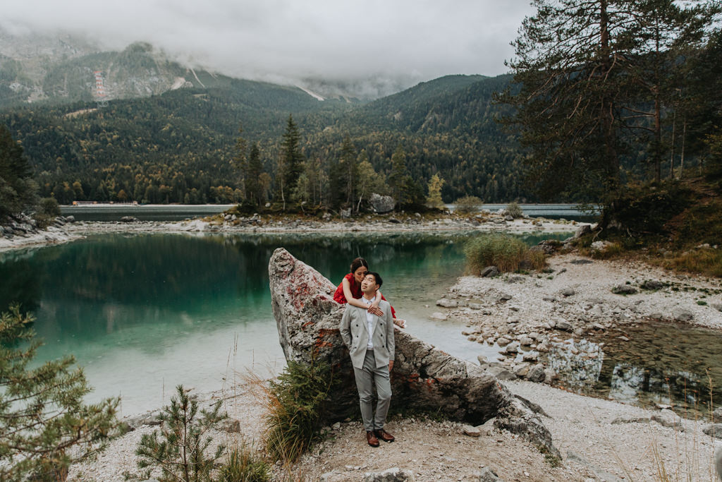 This is a photo of couple eloping at Eibsee during sunrise. They are leaning on a large, triangle shaped boulder. There is a deep green lake surrounding by trees and tall mountain peaks in the background.