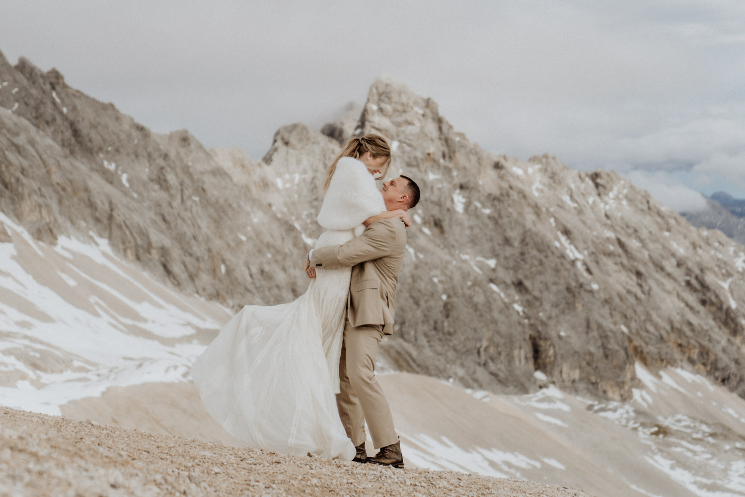 A man holds up a bride in wedding attire while standing on the Zugspitze in Germany. They are facing each other with mountains in the background.