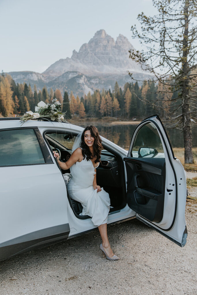 This is an image of a bride stepping out of her rental car during her elopement in Europe. She is wearing a white wedding dress and heels and smiling. 