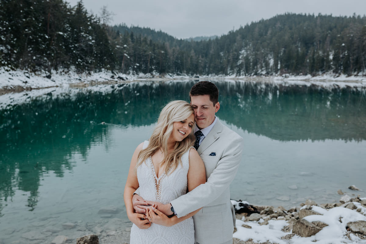 This is an image of a couple in their wedding attire eloping at Eibsee near the Zugspitze. The lake is green blue behind them and there is snow on the trees. They are cuddling with their eyes closed.