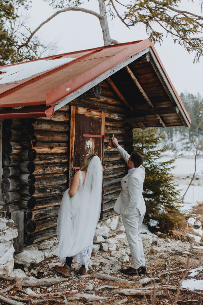 A couple in wedding attire stands outside a mountain hut on their elopement day, pointing to the roof.