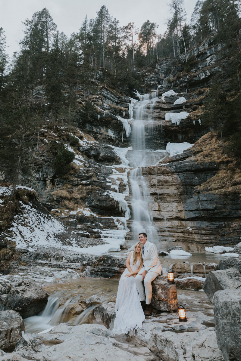 This is a photo of a couple sitting under a snowy waterfall on their elopement day. They are sitting on a large boulder with two warm lanterns by their side. The waterfall is flowing past them and they're staring off to the left of the frame.