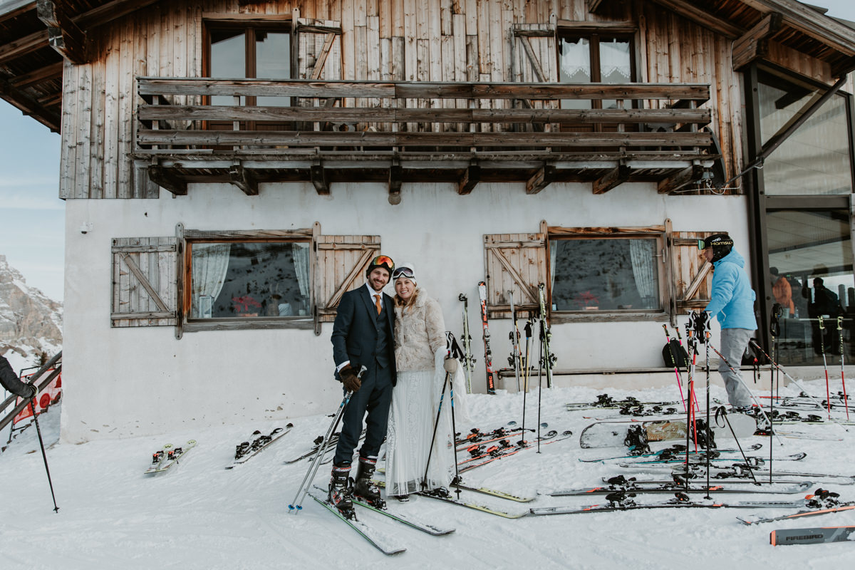 A couple on skis wearing their wedding outfits stands outside a traditional alpine hut in the Dolomites on a sunny winter day.