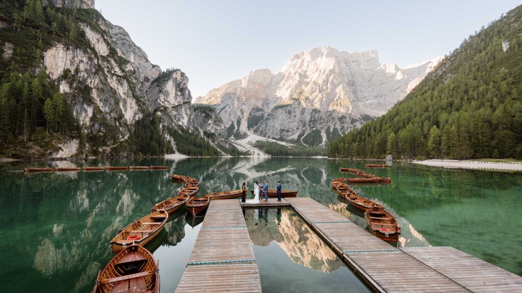A wedding couple stands outside the boathouse at Lago di Braies on their elopement day reading their vows under a clear sunrise sky