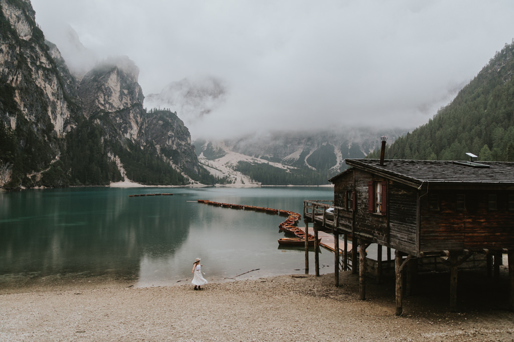 A woman dances in a white dress with the Lago di Braies lake and boat house and misty mountains in the background.