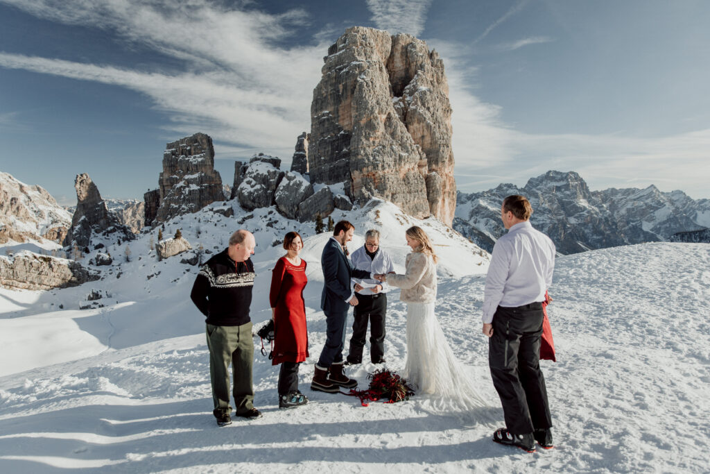 A group of people stand next to a couple exchanging rings on a snowcovered mountain during their minim ony ceremony