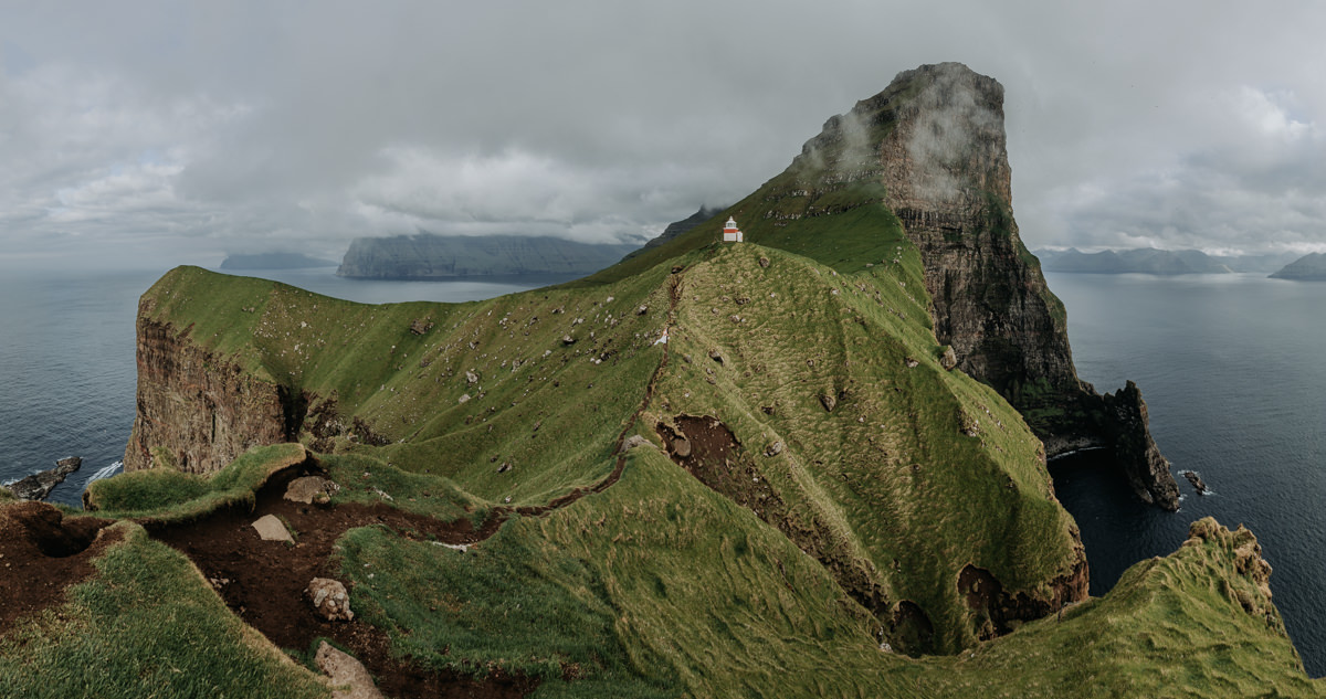 Kalsoy Island in the Faroe Islands, looking back at the lighthouse on a sunny day with clouds covering the tops of the mountains. An eloping couple stands in their wedding attire in the center, as a tiny speck.