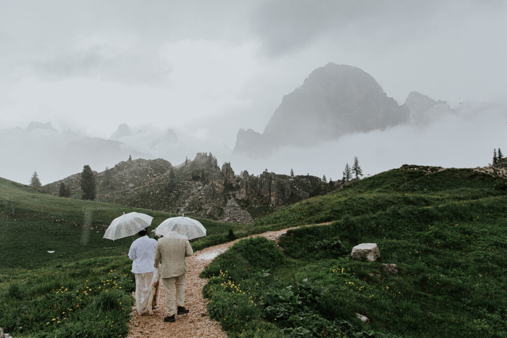 A couple walks in the Dolomites in their wedding attire in the rain, carrying clear umbrellas