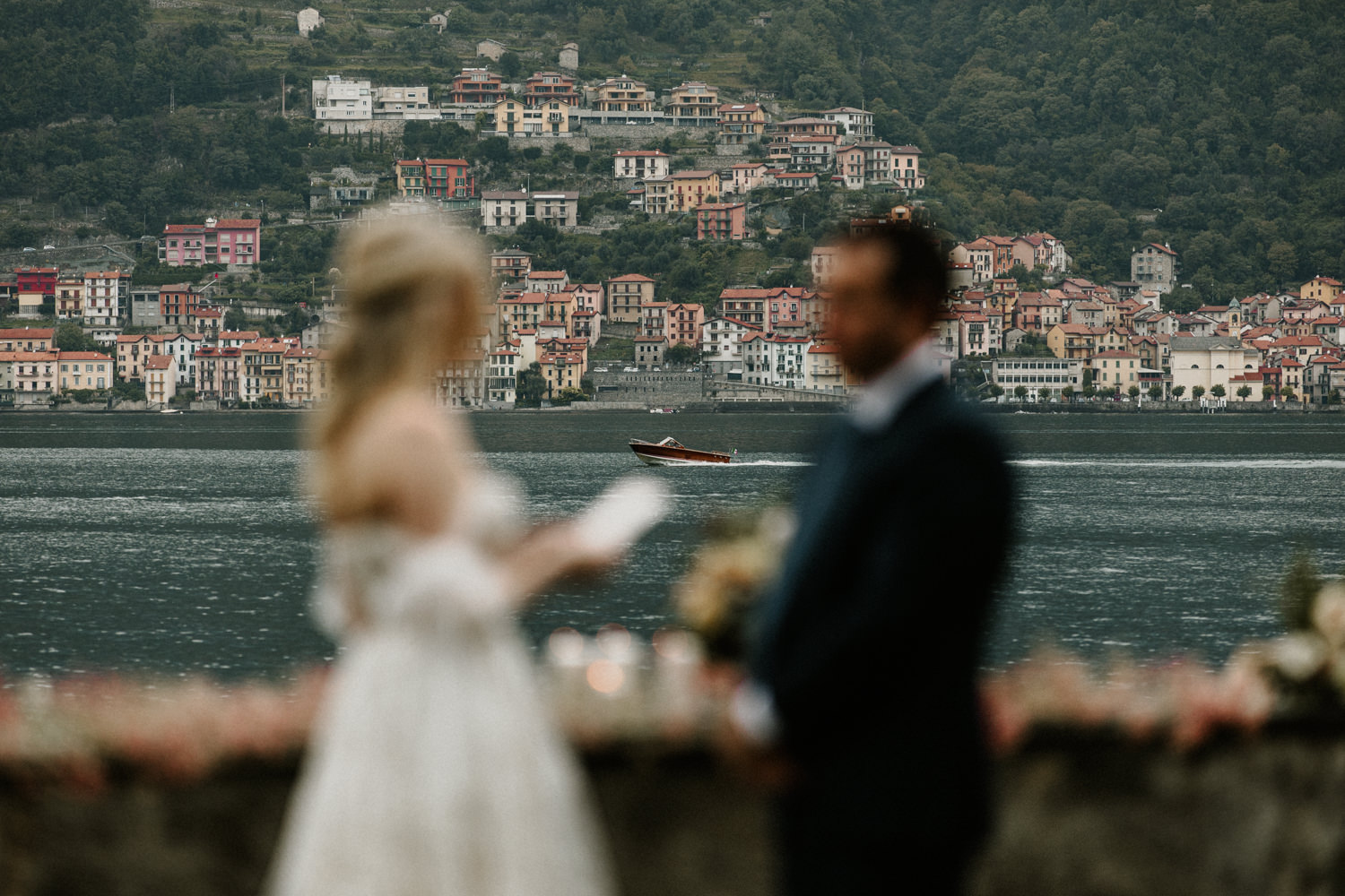 A couple stands out of focus reading wedding vows on the day they elope near Lake Como, Italy. There is a boat in focus on the lake between them, with colorful homes on the hills behind them.
