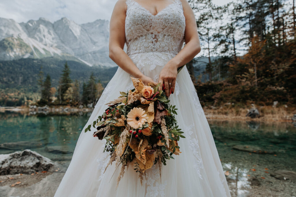 A woman in a wedding gown holds a  bouquet with the Bavarian lake of Eibsee behind her