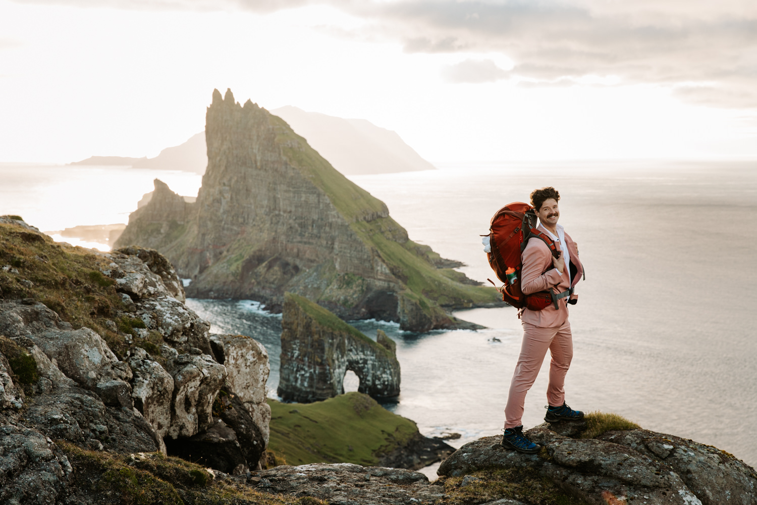 A man with a mustache and pink suit stands near the Drangarnir overlook during his Faroe Islands elopement. He is smiling at the camera and carrying a large orange backpack.